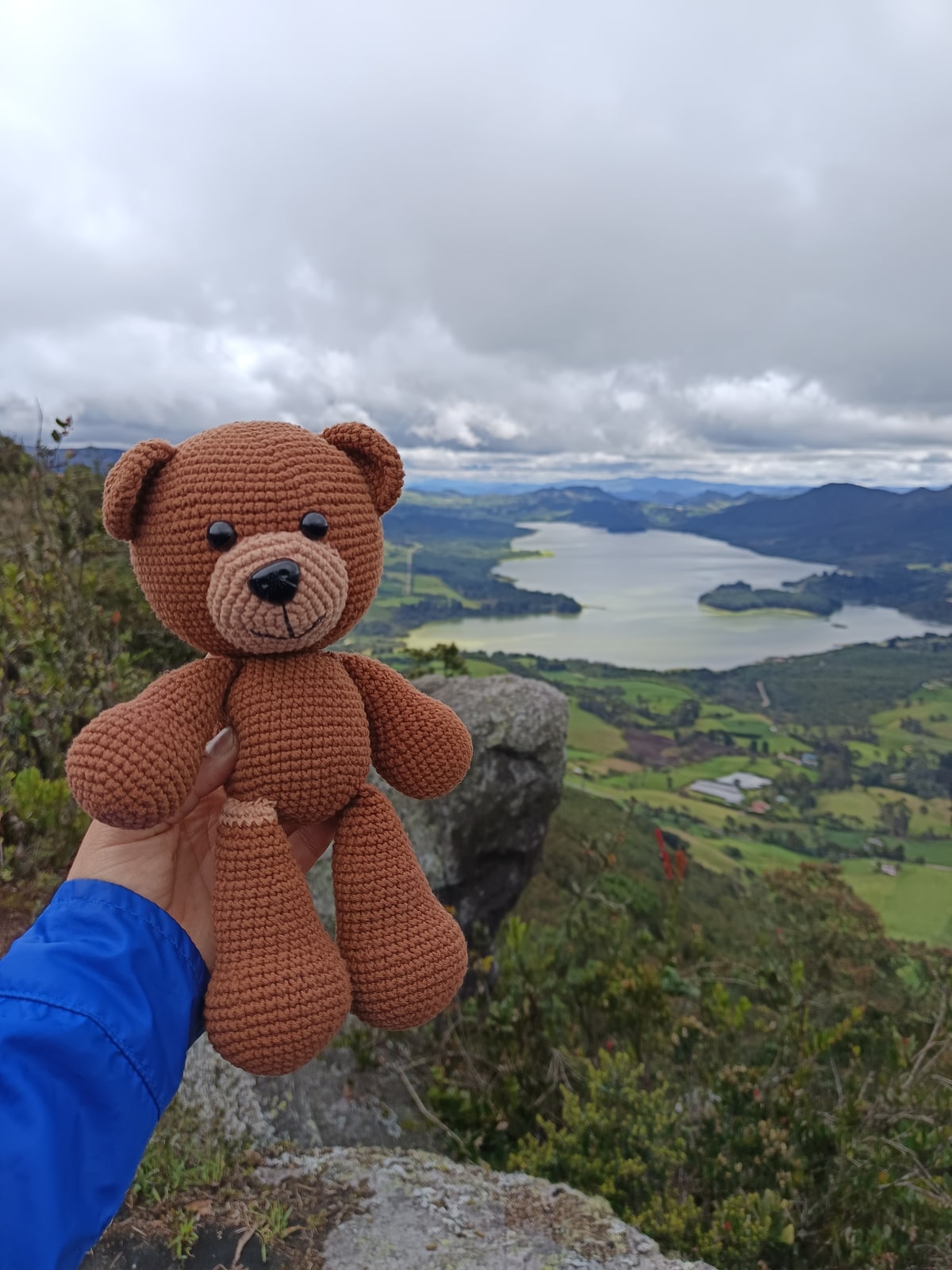 Oso de amigurumi hecho a mano, sostenido frente a un paisaje colombiano